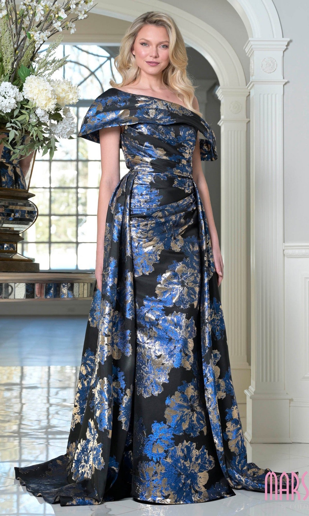 A woman stands indoors in the Marsoni M348 long formal dress, featuring an off-the-shoulder design and floor-length silhouette with royal multi-colored floral jacquard patterns on a black background.
